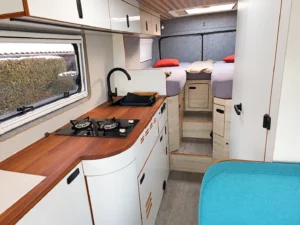 Camperküche mit Holzarbeitsplatte, weißen Möbelfronten und Heckbetten.