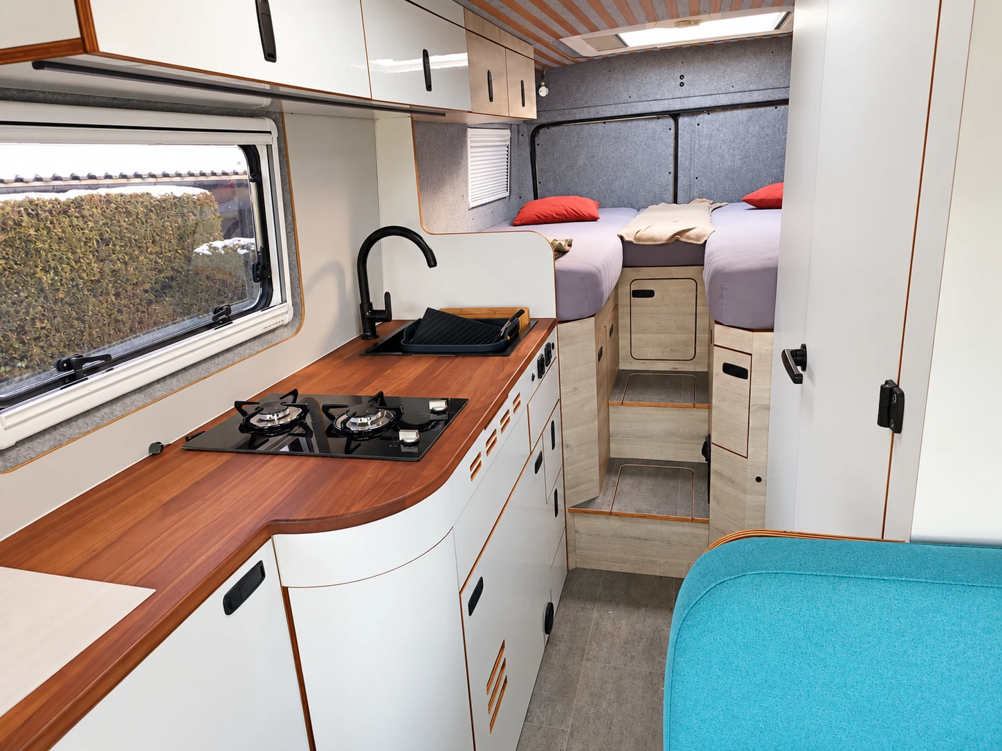 Camperküche mit Holzarbeitsplatte, weißen Möbelfronten und Heckbetten.